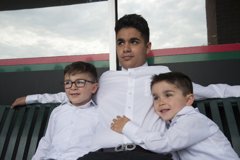 1 teenage boy and 2 smaller boys wearing white shirts, seated on a bench.